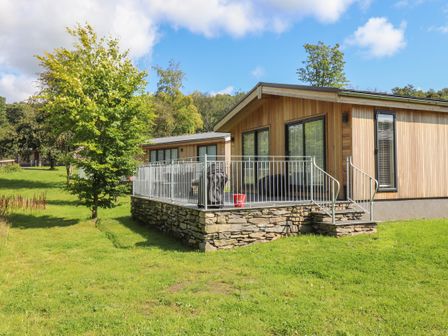 A wooden cabin with a stone base and a metal railing on a grassy lawn with trees at Ash Thwaite in Haverthwaite