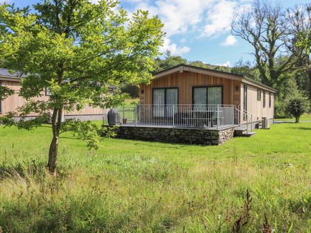 A wooden cabin with patio and railing surrounded by grass and trees at The Brambles in Haverthwaite