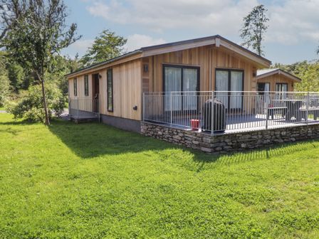 A wooden cabin with a metal railing deck and green lawn surrounding it at The Burrow in Haverthwaite