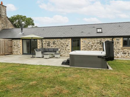 An outdoor patio area with a sofa set and umbrella next to a hot tub in front of a stone building at The Byre in St Brides Major