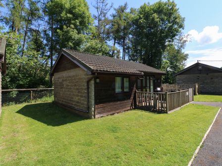 A single story wooden cabin with fenced porch on a grassy area with trees in the background at Chalet Log Cabin L7 in Combe Martin