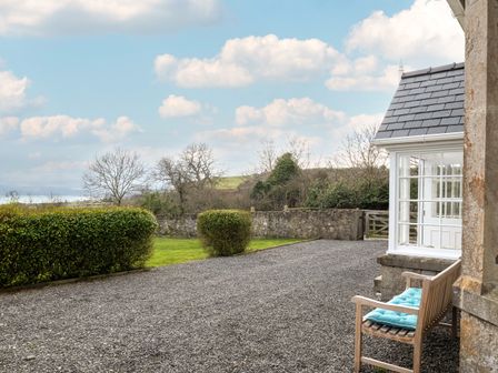 A gravel driveway with a wooden bench and a blue cushion next to a house with white windows and a stone wall at Flagstaff in Penmon near Beaumaris