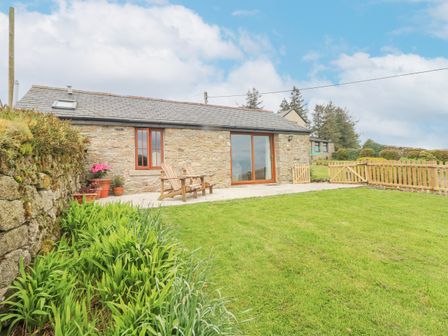 A stone cottage with wooden chairs on a patio and grass lawn surrounded by a wooden fence at Fizz-Gog Cottage in Minions near Upton Cross