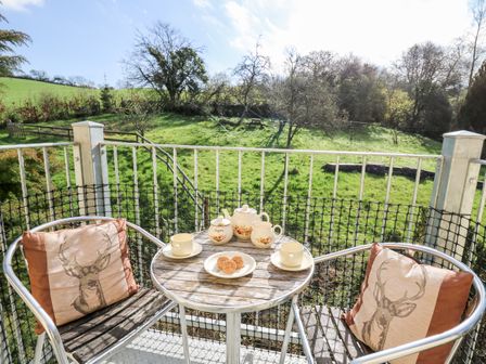 A balcony with a round table set for tea and two chairs with deer cushions overlooking a grassy garden at The Cheese Loft in White Mill near Carmarthen