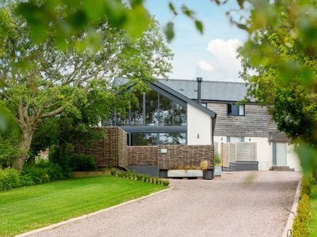 A driveway leading to a house with wooden paneling and large windows surrounded by green trees and grass at The Barn in Llanfechell