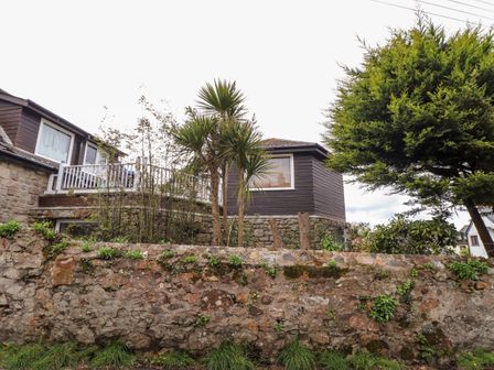 Stone house with wooden upper extension behind a stone wall with plants and trees at The Hexagon House in Goldsithney