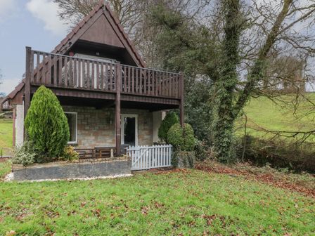 A two-story house with a stone lower floor and wooden balcony surrounded by grass and trees at Valley Lodge 55 Callington near Gunnislake