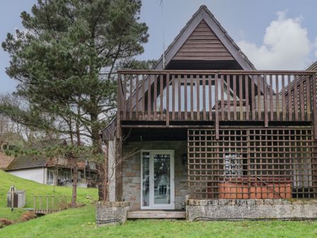 A two-story cabin with a wooden balcony and trellis surrounded by grass and trees at Valley Lodge 33 Callington near Gunnislake