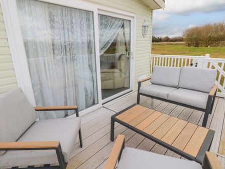 An outdoor deck with cushioned chairs and a wooden table outside a building with sliding glass doors at Park Lane in Llanteg near Kilgetty