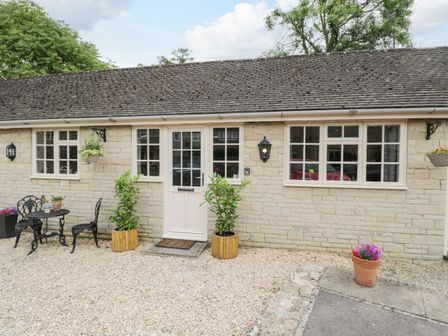 Exterior view of a single-story stone building with a white door and windows with a small metal table and two chairs outside at Well Cottage Nr Malmesbury