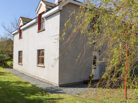 A two-story house with white walls and red window frames surrounded by grass and trees at Wheatfield Lodge in Ramstown near Fethard-On-Sea County Wexford