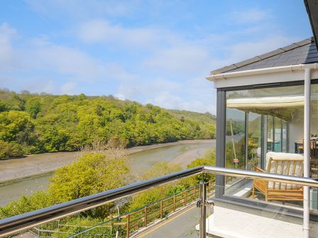 A balcony with glass and metal railing overlooking a river and green trees at Anjoma in Looe