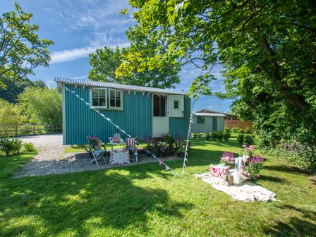 A small blue corrugated metal hut with a picnic table and chairs outside on grass with trees and bunting at Gwithian Shepherds Hut in Angarrack near Hayle