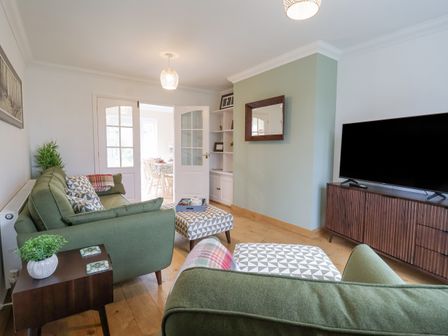 A living room with green sofas a wooden TV stand and a door leading to a dining area at 23 Church Farm Lane in Halesworth