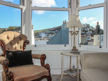 A sitting area with an upholstered armchair a side table with a lamp and a view of houses and water through large windows at 29b Lower Street Dartmouth
