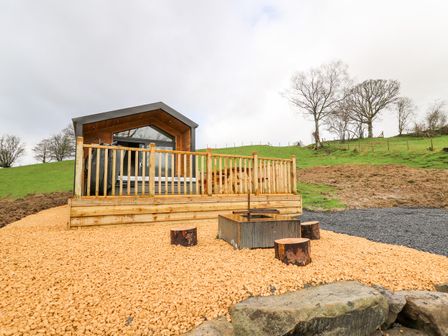 A small wooden cabin with a patio and hot tub on a gravel area with tree stump seats and green grass hill in the background at Fawn's Rest in Nantmel near Rhayader