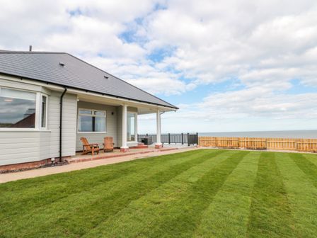 A house with a covered patio and two chairs overlooking a large grass lawn and the sea at Seapoint in Primrose Valley
