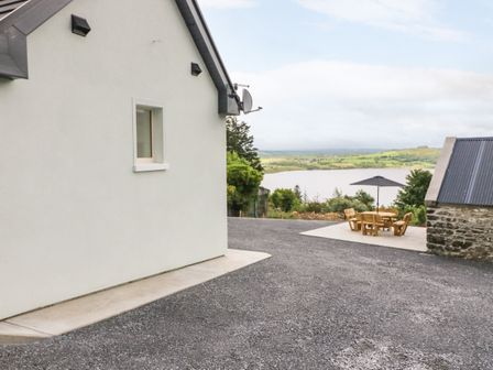 An outdoor area with a white building wall and a small window a stone structure a patio with wooden table and chairs with umbrella and a view of a lake and green hills at An Sean Teach in Muckross near Killarney County Kerry