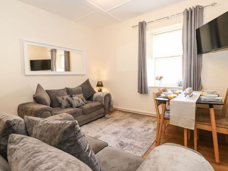 A living room with grey sofas a dining table set with bowls and glasses and a wall mounted TV at The Butcher's Flat in Seahouses