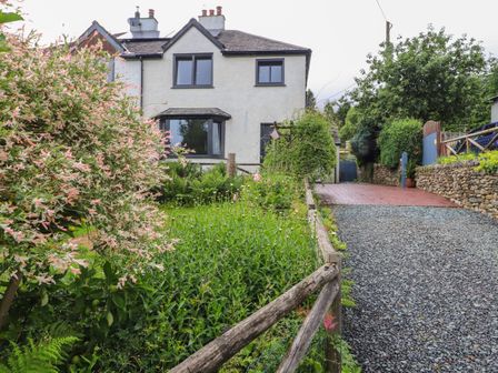 A white two-story house with black window frames surrounded by a garden with plants and a gravel driveway at Beech Croft in Threlkeld near Keswick
