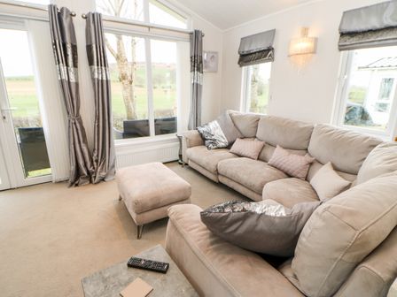 A living room with a beige sectional sofa and ottoman next to large windows with gray curtains at 15 Hesket Caravan Park in High Hesket near Armathwaite