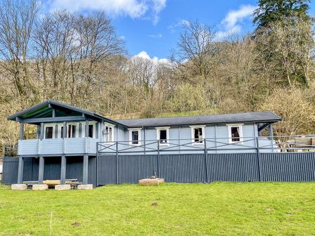 A wooden lodge with a porch surrounded by trees and grass at Valley Lodge in Oakfordbridge near Bampton