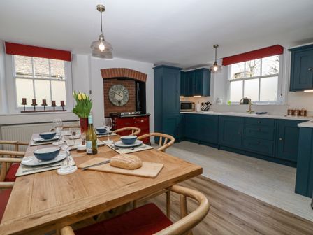 A kitchen with a wooden dining table set with plates bowls wine glasses and a loaf of bread with blue cabinets and two windows at Ellie's Cottage in Burford