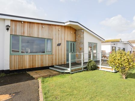 The exterior of a single-story house with wood paneling a green door windows and a small deck with railing at Tinkers Patch in Benllech