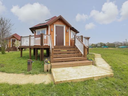 A wooden cabin elevated with stairs and railings on a grassy field at Dragonfly Retreat in Upper Marham