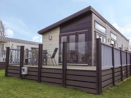 A modern exterior of a lodge with glass and wood railing on a raised deck with outdoor chairs and table at Tiptoes Lodge in Carnforth