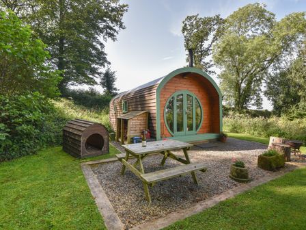 A wooden curved cabin with green framed round doors and a picnic table on gravel at Pendragon Pod in Axminster