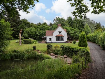 A white house with a red roof surrounded by greenery and a pond in front at Grove Cottage in Leominster