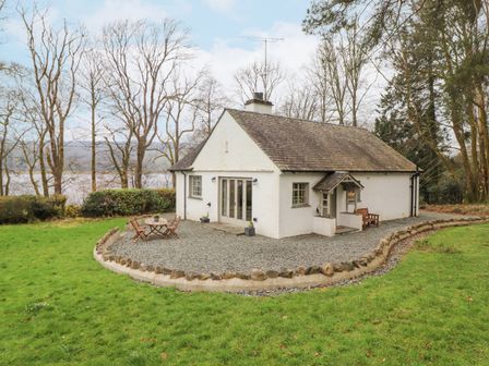 A white cottage with a stone patio and outdoor table and chairs surrounded by grass and trees at Larch Cottage at Esthwaite Water in Hawkshead