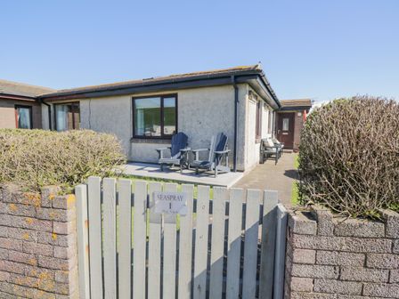 A front view of a single-story house with two chairs on the patio behind a wooden gate at Seaspray in St Bees