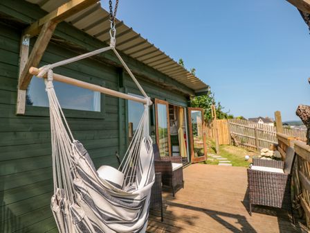 An outdoor wooden deck with a hanging chair a white hat on the chair and wicker chairs along a green wooden building at Dart View Hideout in Totnes
