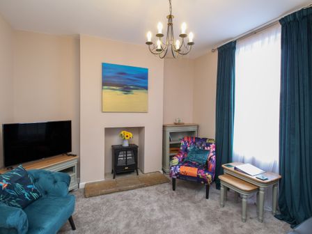 A living room with a blue armchair a colorful armchair a television a small fireplace and blue curtains at The Skipper's House in Weymouth