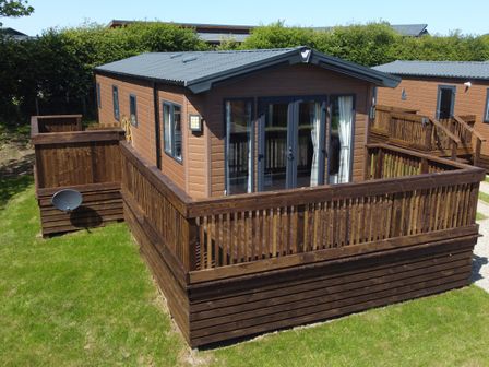 A wooden cabin with a fenced deck and glass doors on a grassy area at Rose Lodge in Runswick Bay near Staithes