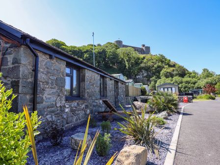 Stone building with windows and plants in front near a paved road with a castle on a hill in the background at Tan Llech in Harlech