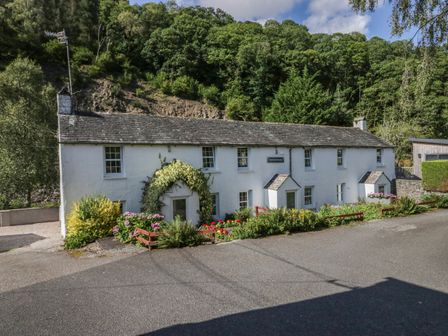 A white cottage with multiple windows and flower beds in front surrounded by trees at Riverside Cottage No 3 Keswick