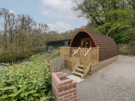 A small wooden pod with a front porch near a river and a green metal bridge in a wooded area at Pod No 5 in Keswick