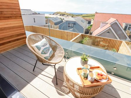 A balcony with a round chair with a fish pillow and a round table with cups and snacks overlooking houses and the sea at 11 Ebbtide in Porth