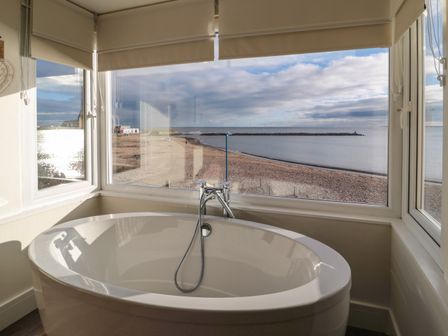 A bathroom with a bathtub near large windows overlooking a beach and sea at Fairwinds in Newbiggin-By-The-Sea