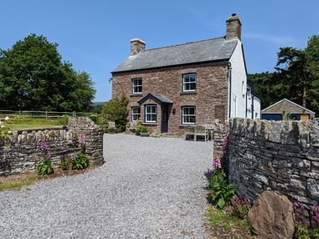 A stone house with a gravel driveway and wooden outdoor furniture surrounded by stone walls and flowers at Ty Sinsir in Tretower