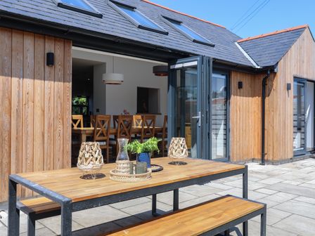 An outdoor patio with a wooden table and benches next to a house with sliding glass doors at White Lodge Cottage in Niwbwrch newborough