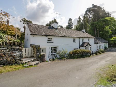 A white cottage with a slate roof beside a paved road surrounded by stone walls and trees at Old Farm Cottage in Skelwith Fold
