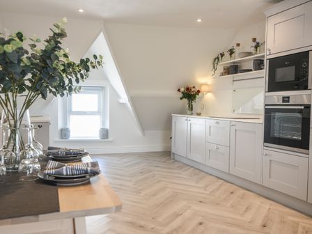 A kitchen with white cabinets a built in oven and microwave a wooden dining table with place settings and plants at Fore Street House - Apartment 3 in Seaton