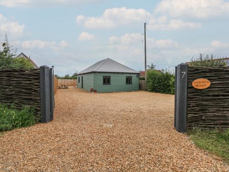 A gravel driveway leading to a green house with a metal roof behind a woven fence at Tawny's Roost in Snettisham