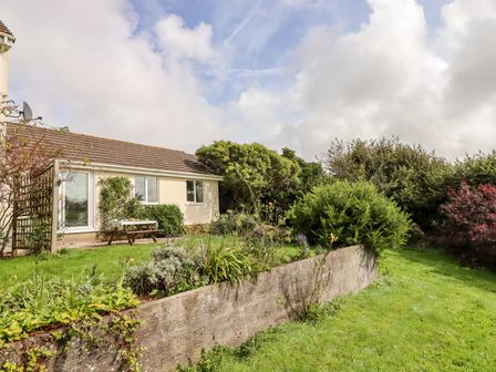 A garden with grass shrubs and flowering plants beside a single-story cream house with two windows and a picnic bench at Mabel's View in East Allington Devon
