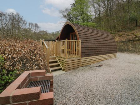A wooden pod with a small porch and chairs surrounded by trees and a gravel ground at Pod No 6 in Keswick