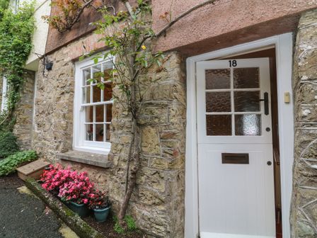 A stone exterior wall with a white door numbered 18 and a window next to flower pots with pink flowers at 18 Robinsons Row in Salcombe